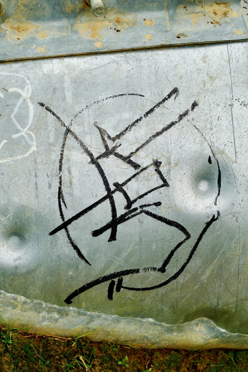 Another tag, black paint pen in a circle with square linear glyphs in inside. It's on a metal plate.