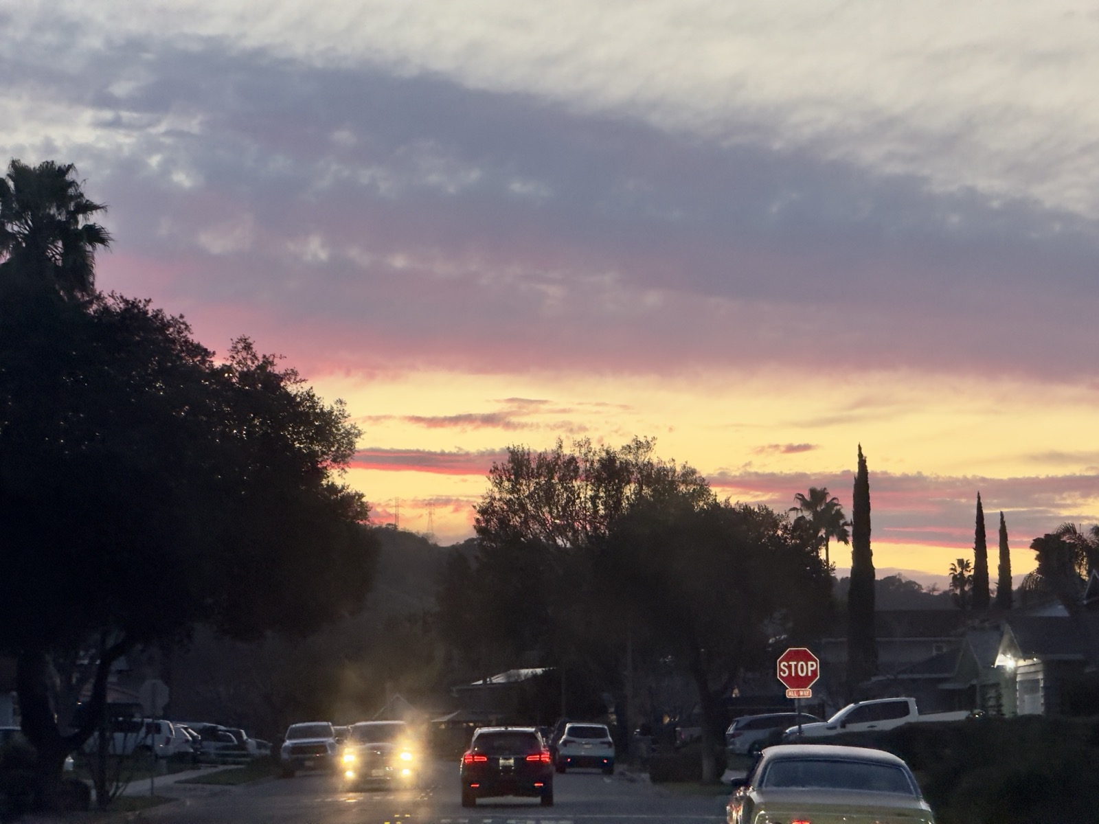 A sunset in pink, purple, lavender, yellow, and blue. It was just a corner of the sky, but an electric corner. Below the sky, a few cars and a stop sign.