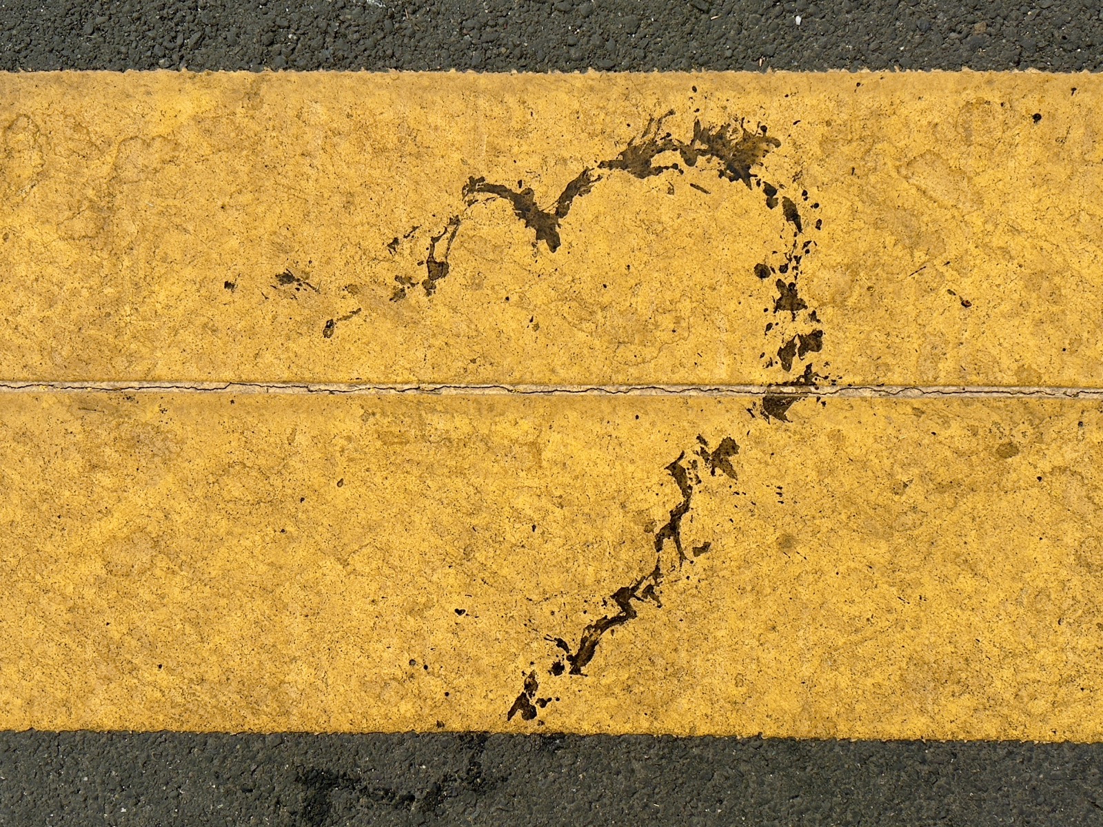 There is a wide yellow paint stripe from a crosswalk, and over it there is a dark dripped greasy line that is evocative of a drawn heart.