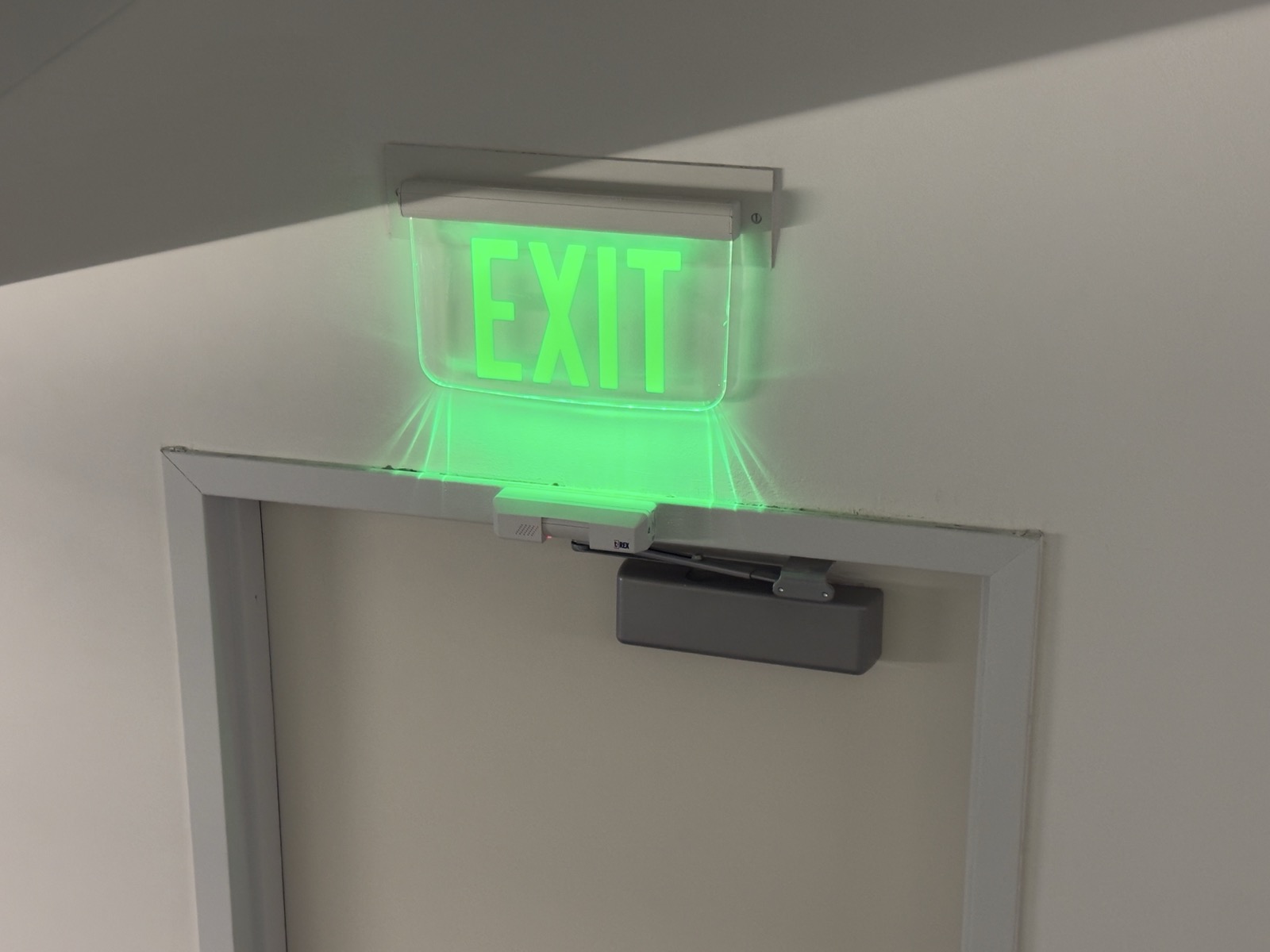 A green illuminated exit sign, with green light spilling out of the bottom, above a door.